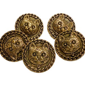 May include: Five antique gold-colored buttons with embossed cat faces. Each button features a detailed cat portrait within a decorative rope-like border. The buttons are round and have a vintage aesthetic.