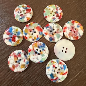 Op de afbeelding: Een verzameling van tien ronde knopen. Negen knopen hebben een mozaïekontwerp met rode, blauwe, gele en groene fragmenten op een witte achtergrond. Eén knoop is effen wit. Elke knoop heeft vier gaten om te naaien.