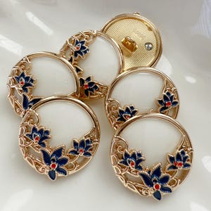 May include: A collection of decorative buttons with a gold-tone metal frame. Each button features a white center and is adorned with blue floral designs and red accents. The buttons have a shank back.