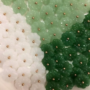 May include: A collection of small, flower-shaped beads in shades of white, light green, and dark green. Each bead features a smooth, carved petal design with a small, metallic gold-toned center accent, arranged in a dense, overlapping pattern.