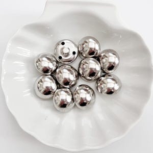 Puede incluir: Una colecci&oacute;n de broches met&aacute;licos plateados y brillantes. Los broches tienen forma de c&uacute;pula y est&aacute;n dispuestos en un plato blanco con volantes. Un broche tiene una base con p&uacute;as visible, los otros son lisos y redondeados.