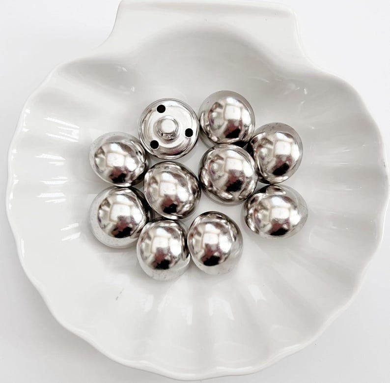 Puede incluir: Una colecci&oacute;n de broches met&aacute;licos plateados y brillantes. Los broches tienen forma de c&uacute;pula y est&aacute;n dispuestos en un plato blanco con volantes. Un broche tiene una base con p&uacute;as visible, los otros son lisos y redondeados.