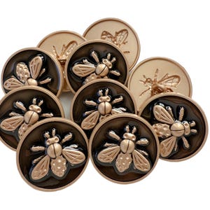 May include: A collection of bronze-colored buttons with a bee design. The buttons have a dark brown background and a raised bee motif. Some buttons have a smooth finish, while others have a textured appearance. The buttons are round and vary in size.
