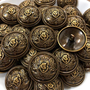 May include: A collection of round, antique bronze-coloured metal buttons featuring a raised relief design of a shield with the letter D, flanked by two lions and topped with a crown. Each button has a circular border and a shank on the back.