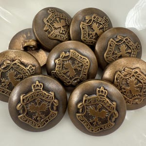 May include: A collection of antique bronze-colored buttons. Each button features a detailed crest design with a crown and a bird emblem. The buttons are round and have a slightly domed shape, suggesting a vintage or historical theme.