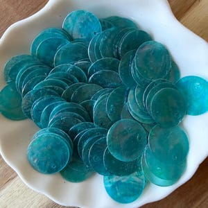 May include: A collection of circular, teal-colored shell sequins with an iridescent finish. Each flat, round piece features a small, off-center hole for attachment, showcasing natural variations in the blue-green surface texture and luster.