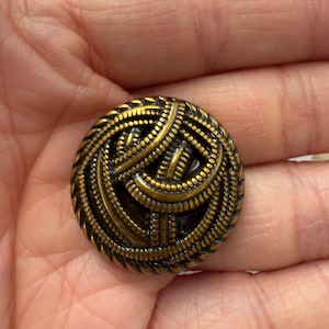 May include: A round, antique brass button with a detailed, woven design. The button has a dark, aged finish with intricate patterns and a textured surface. The button's design features a series of interwoven lines and small, raised dots.