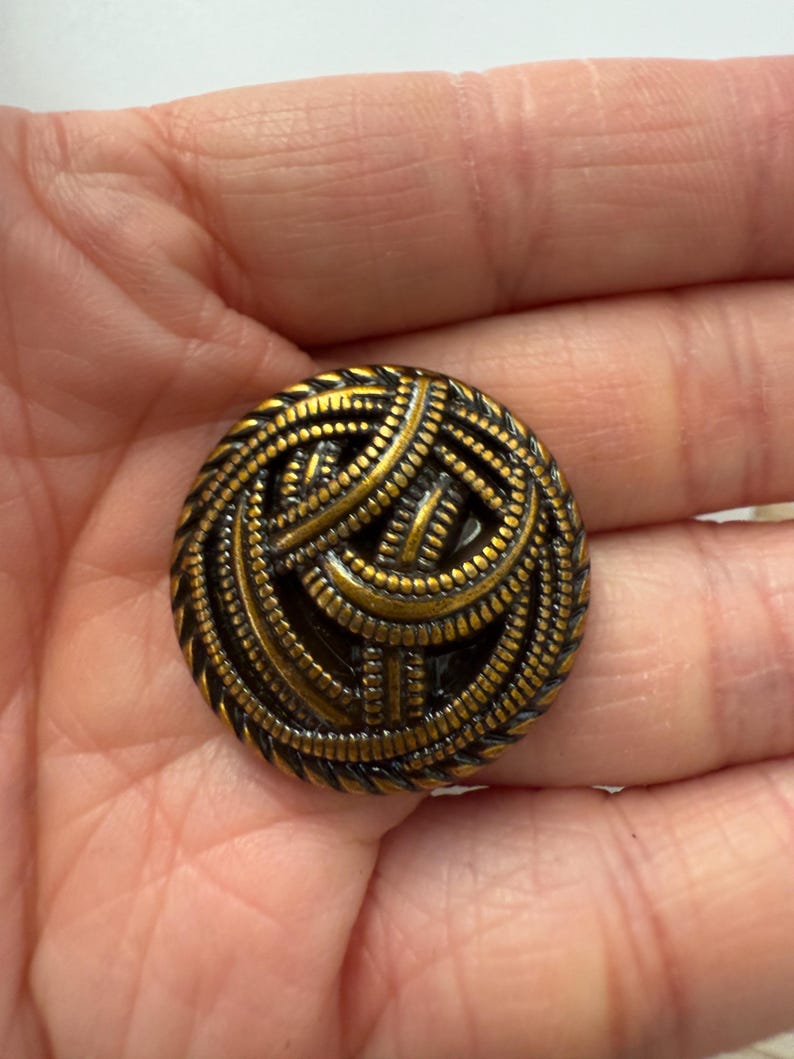 May include: A round, antique brass button with a detailed, woven design. The button has a dark, aged finish with intricate patterns and a textured surface. The button's design features a series of interwoven lines and small, raised dots.