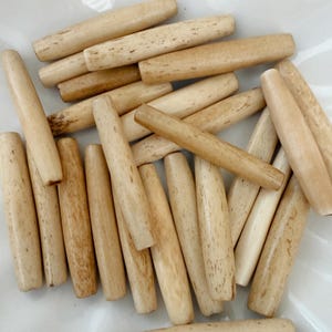 May include: A collection of light-colored wooden beads, each cylindrical with rounded ends. The beads have a natural wood grain pattern and vary slightly in color, ranging from cream to light brown. They are scattered on a white surface.