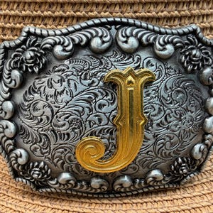 May include: A silver-colored, ornate belt buckle with a gold-colored letter "J" in the center. The buckle has a detailed floral and scroll design. The buckle is set against a straw hat with a black band.