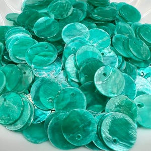 Puede incluir: Una gran colección de discos circulares de concha en color verde azulado. Cada pieza plana y redonda presenta una superficie lisa e iridiscente con un brillo nacarado natural y un pequeño orificio perforado cerca del borde superior.