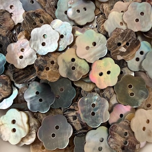 May include: A collection of flower-shaped buttons crafted from iridescent mother-of-pearl shell. Each button features a scalloped edge and two central holes for attachment, displaying a variety of natural tones including brown, gray, and cream.
