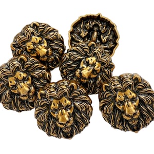 May include: A selection of antique brass-coloured buttons, each displaying a detailed lion's head design. The buttons have a textured finish and are arranged on a white background. The lion's mane and facial features are intricately sculpted.