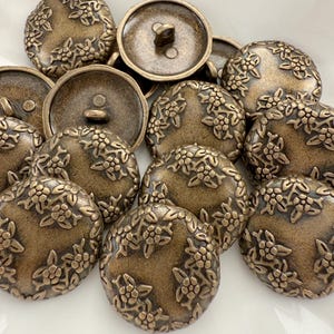 Può includere: Una collezione di bottoni antichi color bronzo. Ogni bottone è rotondo con un motivo floreale sui bordi e un occhiello sul retro per cucire. I bottoni hanno un diametro di circa 2,5 cm.