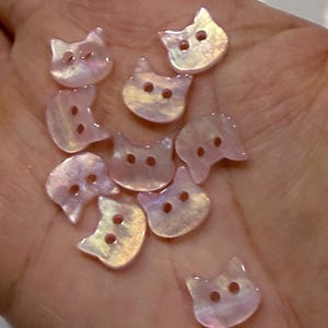 Puede incluir: Un conjunto de botones iridiscentes con forma de gato. Cada botón tiene dos pequeños agujeros para coser y un acabado nacarado que refleja la luz, creando un efecto brillante. Los botones son de color rosa pálido.