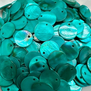 Puede incluir: Una colección de discos circulares de concha de color turquesa, cada uno con un pequeño orificio cerca del borde superior. Las superficies exhiben un brillo iridiscente natural con texturas y profundidades de color variadas y distintivas.