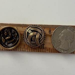 May include: Two vintage brass buttons with intricate designs, one featuring an anchor and the other a woven pattern. A quarter coin is included for size reference. The buttons are approximately 1 inch in diameter, perfect for sewing projects.