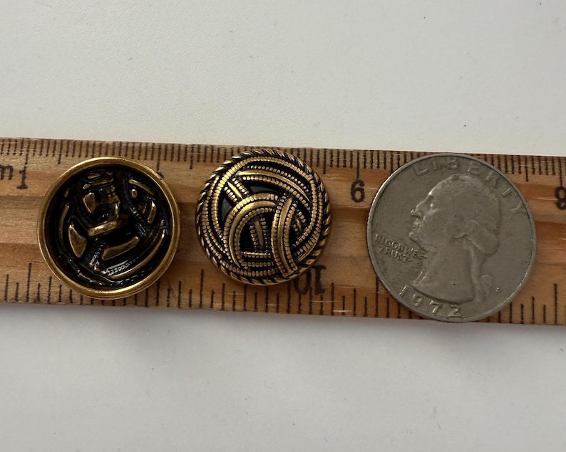 May include: Two vintage brass buttons with intricate designs, one featuring an anchor and the other a woven pattern. A quarter coin is included for size reference. The buttons are approximately 1 inch in diameter, perfect for sewing projects.