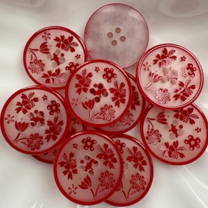 Puede incluir: Un conjunto de botones redondos con un diseño floral rojo. Los botones tienen un centro blanco translúcido y un borde rojo. Algunos botones tienen cuatro agujeros para coser.