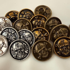 May include: A collection of round metal buttons featuring a raised skull and crossbones design. The buttons are finished in various metallic tones, including silver, antique gold, and bronze, with detailed engravings on each skull's forehead.