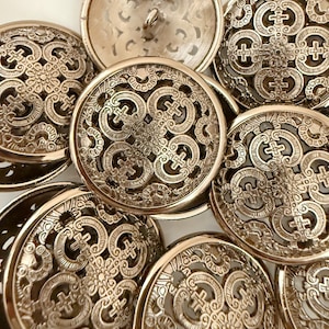 Può includere: Una collezione di bottoni metallici ornati con intricati disegni traforati. I bottoni sono rotondi con un motivo in rilievo con croci ed elementi floreali. Il colore è un oro o bronzo tenue, e i bottoni sono disposti in una ripresa ravvicinata.