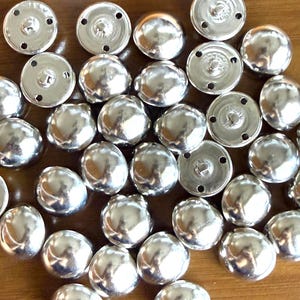 Puede incluir: Una colecci&oacute;n de botones de presi&oacute;n de metal plateado brillante. Los botones tienen una parte superior redondeada y abovedada y una base plana y circular con agujeros para coser. Los botones est&aacute;n dispersos sobre una superficie de madera.