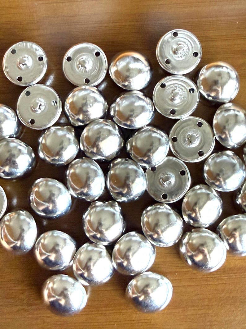 Puede incluir: Una colecci&oacute;n de botones de presi&oacute;n de metal plateado brillante. Los botones tienen una parte superior redondeada y abovedada y una base plana y circular con agujeros para coser. Los botones est&aacute;n dispersos sobre una superficie de madera.