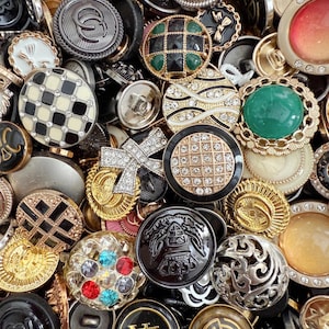 May include: A collection of various buttons in different shapes, sizes, and colors. The buttons include gold, silver, black, and white, with embellishments like rhinestones, enamel, and intricate designs. Some buttons have a checkerboard pattern, while others feature geometric shapes.