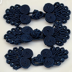 May include: Three dark navy blue frog closures made of coiled cord and fabric knots. Each fastener features a symmetrical design with two large central spirals and decorative loops on the outer edges, accented with small matching beads.