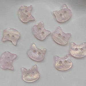 May include: A collection of ten iridescent pink cat-shaped buttons. Each button has two holes for sewing. The buttons are made of a translucent material with a pearlescent sheen, creating a subtle, shimmering effect. Ideal for craft projects.