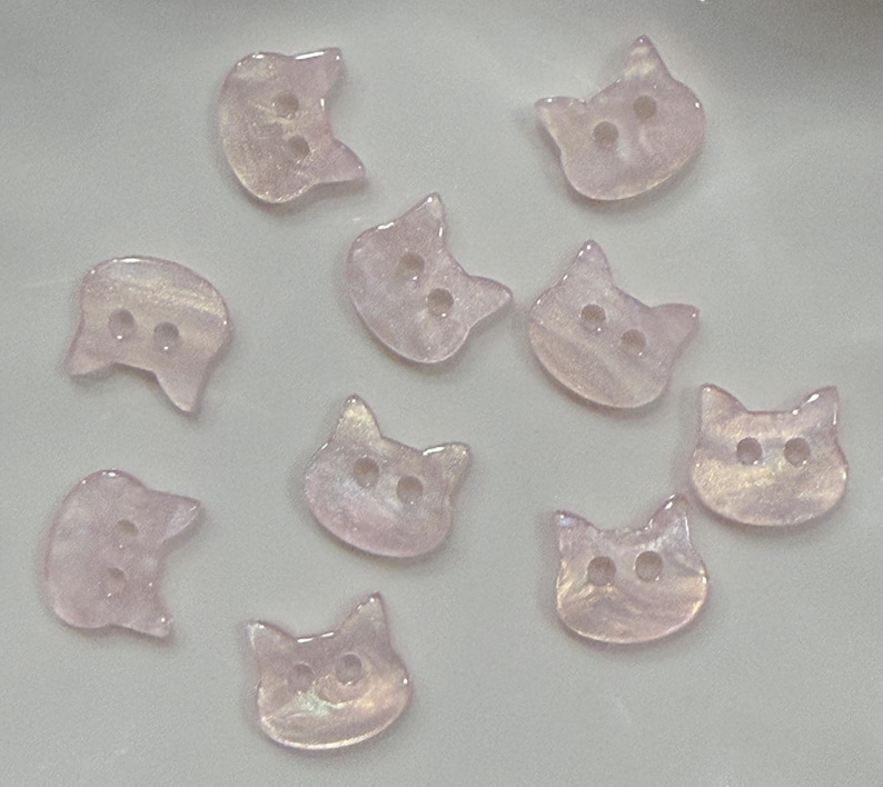May include: A collection of ten iridescent pink cat-shaped buttons. Each button has two holes for sewing. The buttons are made of a translucent material with a pearlescent sheen, creating a subtle, shimmering effect. Ideal for craft projects.