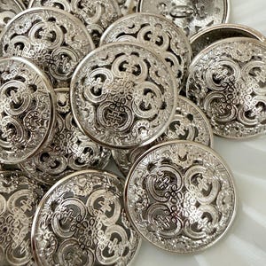 May include: A collection of silver-colored, ornate buttons. Each button has a detailed, openwork design featuring swirling patterns and a central cross-like motif. The buttons are round and have a shiny, metallic finish, suggesting they are made of metal.
