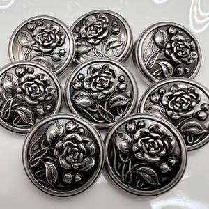 May include: Nine antique silver-toned buttons with a raised rose and leaf design. The buttons are round and have a detailed floral pattern. The buttons are likely made of metal.