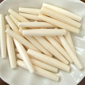 May include: A white, shallow, scalloped-edge plate filled with numerous off-white, cylindrical food items. The food items are arranged in a random pattern, with some overlapping. The plate is set against a neutral background.