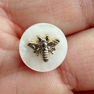 May include: A round, white button featuring a gold-coloured bee motif. The bee has intricate wing and body detailing. The button is held in a person's hand.