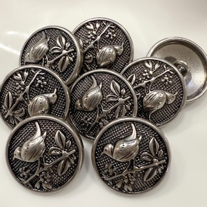 May include: A collection of antique silver-toned metal buttons. Each button features a detailed relief design of a bird perched on a branch with leaves and berries. The buttons are round and have a slightly textured surface.