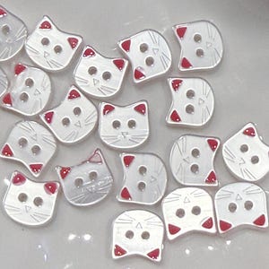 May include: A collection of white cat-shaped buttons with red accents on the ears. Each button has two or three holes for sewing. The buttons are made of a shiny, smooth material and are arranged on a light-colored surface.