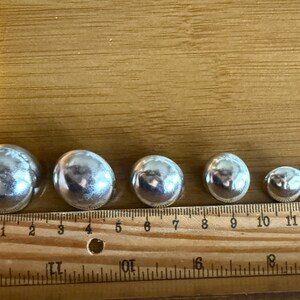 Puede incluir: Una colecci&oacute;n de siete esferas plateadas brillantes de diferentes tama&ntilde;os, dispuestas en fila. Una regla de madera con marcas de cent&iacute;metros se coloca junto a las esferas para referencia de tama&ntilde;o. Las esferas var&iacute;an de 1,3 cm a 3,2 cm de di&aacute;metro.