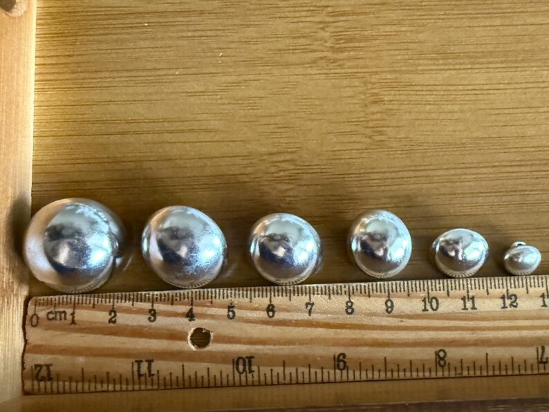 Puede incluir: Una colecci&oacute;n de siete esferas plateadas brillantes de diferentes tama&ntilde;os, dispuestas en fila. Una regla de madera con marcas de cent&iacute;metros se coloca junto a las esferas para referencia de tama&ntilde;o. Las esferas var&iacute;an de 1,3 cm a 3,2 cm de di&aacute;metro.