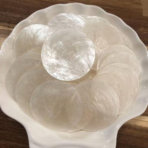 May include: A collection of circular, translucent white mother-of-pearl shell discs with a natural iridescent sheen. Each flat, thin disc features a smooth surface and a small pre-drilled hole near the edge, arranged in a white scalloped bowl.