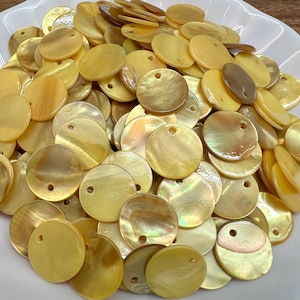 Peut inclure: Une grande collection de boutons circulaires en nacre dans des tons jaune pâle et crème. Chaque bouton présente une surface lisse et irisée avec un petit trou près du bord supérieur pour la fixation, mettant en valeur les variations naturelles.