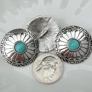May include: Three silver-toned metal buttons with a turquoise center stone. The buttons have a detailed geometric design around the edges. A quarter coin is included for size comparison. The buttons are likely for clothing or craft projects.