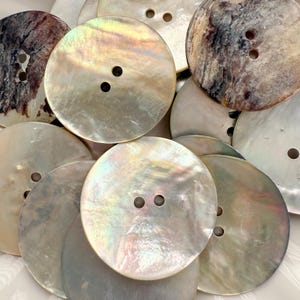 May include: A collection of large, round mother-of-pearl buttons. The buttons vary in color, with iridescent, cream, and brown tones. Each button has two holes for sewing. The buttons are arranged in a pile.