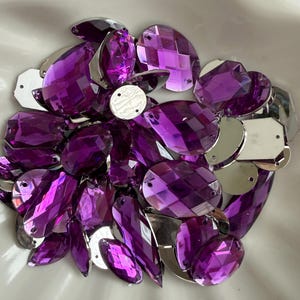 May include: A collection of faceted, purple crystal rhinestones with silver metal settings. The rhinestones are various shapes, including oval, teardrop, and rectangular. The crystals are arranged in a white bowl.
