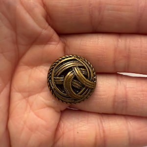 May include: A round, antique brass button with a woven design. The button features a detailed pattern of interwoven lines and a textured edge. The button is a vintage style and is likely used for clothing or crafts.