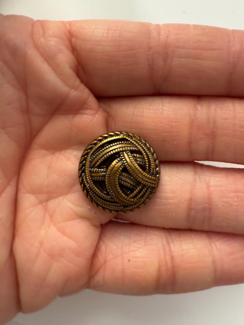 May include: A round, antique brass button with a woven design. The button features a detailed pattern of interwoven lines and a textured edge. The button is a vintage style and is likely used for clothing or crafts.