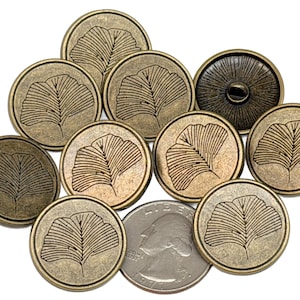 May include: A collection of antique brass-colored buttons with a leaf design. The buttons are round and feature a detailed illustration of a ginkgo leaf. A US quarter is included for size comparison. The buttons are likely for sewing or crafts.