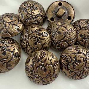 May include: A collection of antique bronze-colored buttons with an ornate, swirling floral design. The buttons are round and domed, with a detailed pattern. One button has a shank for sewing. The buttons are likely for clothing or craft projects.