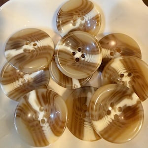 May include: A collection of round, four-hole buttons featuring a swirling, marbled pattern of translucent beige and opaque white tones. Each button has a smooth, glossy surface with a distinct diagonal band of white running through the center.