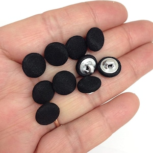 May include: A collection of black fabric-covered buttons with silver metal backs. The buttons are round and have a textured surface. These buttons are suitable for sewing projects and garment embellishment.